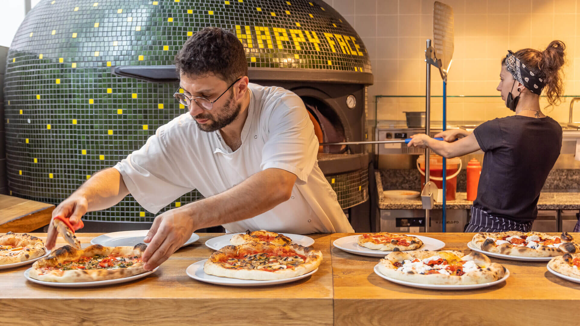 People making pizzas in front of an old-fashioned wood fired oven at Happy Face. There are eight pizzas visible on a wooden counter.
