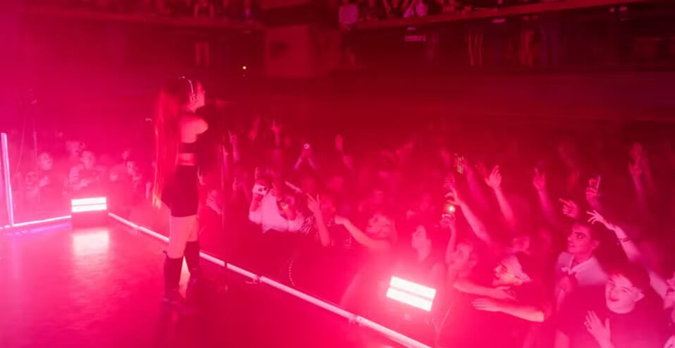 Inside of Scala, with a performer on stage surrounded by fans at ground and elevated levels, and pink light filling the stage.