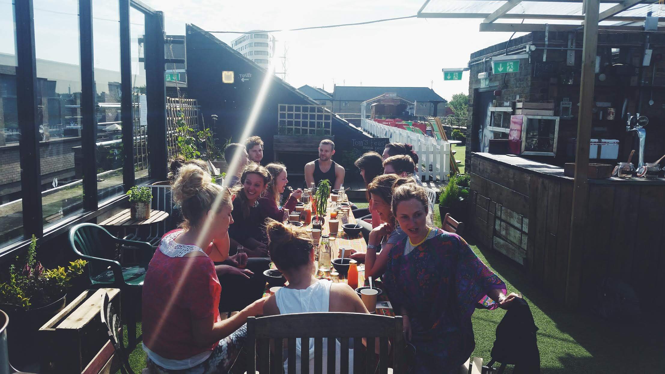 A rooftop bar in Hoxton, by the railway. Atop it is wooden furniture, a play area for children, and a long table with friends gathered about.