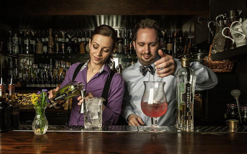Two bartenders at The Nightjar mixing drinks in front of an old fashioned bar with lots of drink options.
