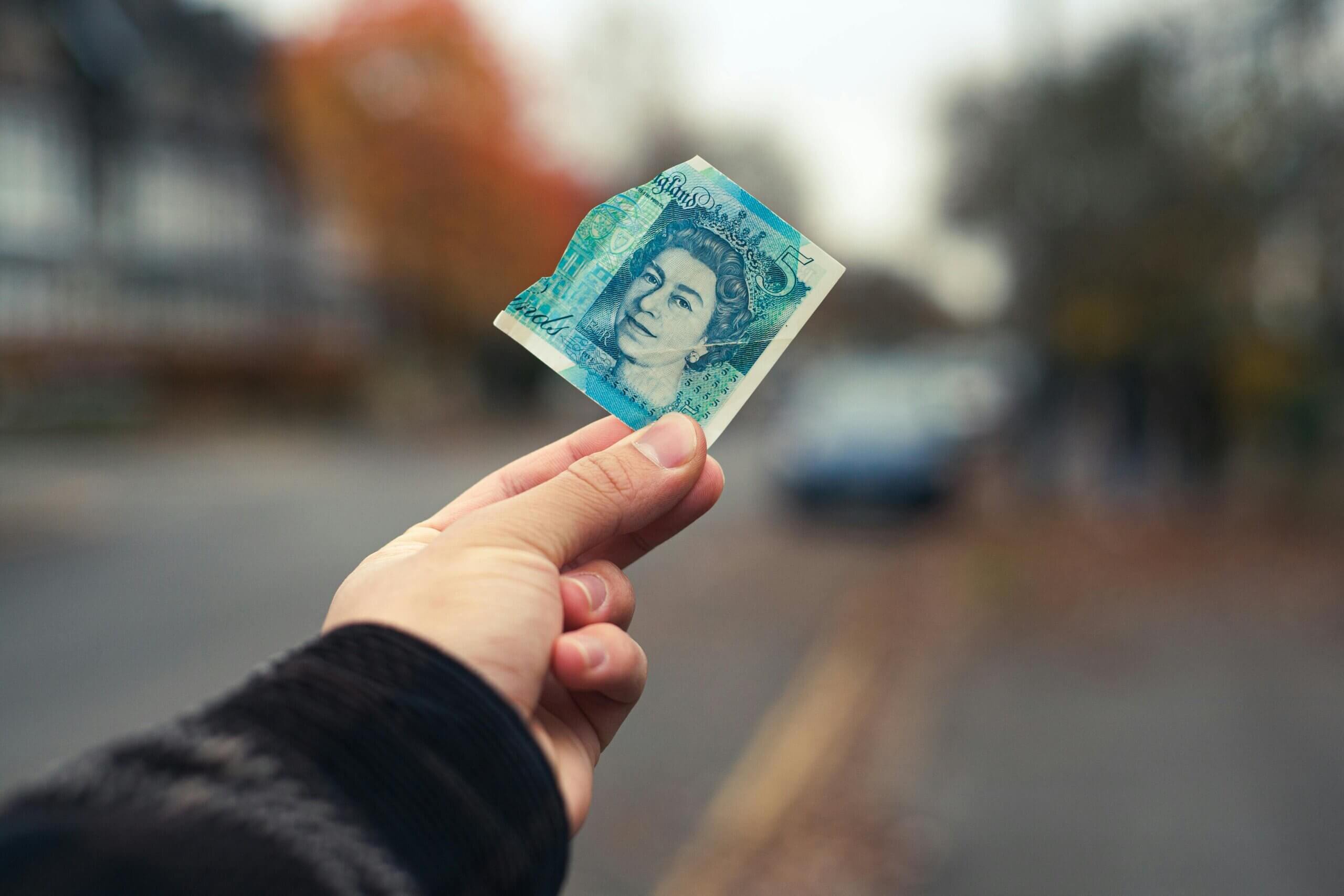 Someone holds a £5 out between a finger and a thumb in the street.