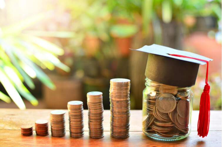 towers of coins lead towards a jar filled with coins, topped with a graduation cap. The background is a green garden and bricks. 
